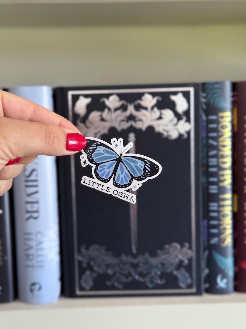 K&ouml;nnte beinhalten: Ein Aufkleber mit einem blau-schwarzen Schmetterlingsdesign und dem Text "LITTLE OSHA". Der Aufkleber wird vor einem dunkelblauen Buch mit silbernen floralen Akzenten und anderen B&uuml;chern auf einem Regal gehalten.