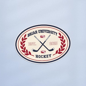 May include: Oval sticker with a cream background featuring the text "Briar University Hockey" and crossed hockey sticks. The design includes the text "Est. 2015" and a red and blue colour scheme. A red laurel wreath frames the design.