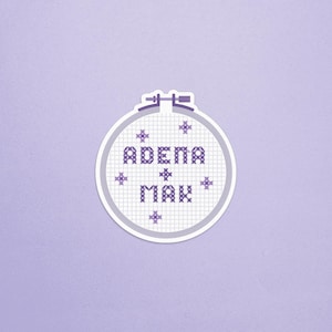 May include: A white embroidery hoop with purple thread, featuring the names "ADEMA" and "MAX" in a cross-stitch pattern.