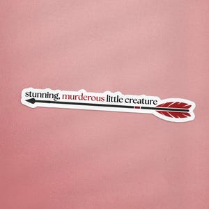 Op de afbeelding: Een witte sticker in de vorm van een pijl met de tekst "stunning, murderous little creature". De pijl heeft een zwarte schacht, een zwarte pijlpunt en rode veren. De achtergrond is effen roze.