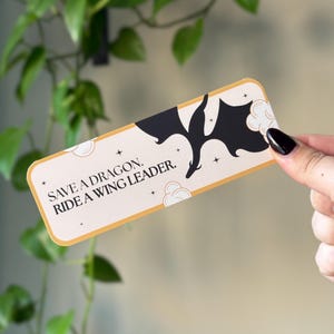 May include: A gold and white bookmark with a black silhouette of a dragon flying through clouds. The bookmark has the text "Save a dragon, ride a wing leader."