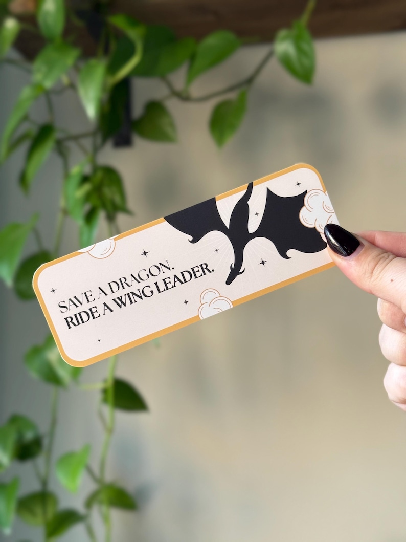 May include: A gold and white bookmark with a black silhouette of a dragon flying through clouds. The bookmark has the text "Save a dragon, ride a wing leader."