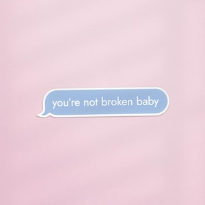 May include: A light blue sticker in the shape of a speech bubble with the text "you're not broken baby" in white. The sticker is set against a soft pink background.