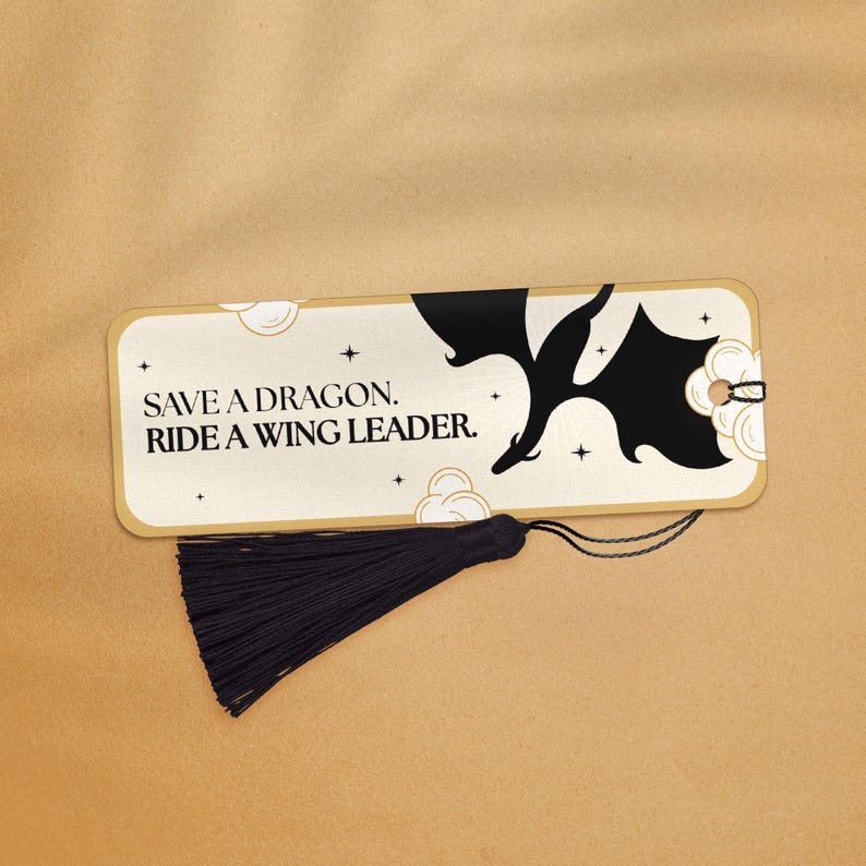 May include: Gold bookmark with a black silhouette of a dragon flying with the text "Save a dragon. Ride a wing leader." and a black tassel.