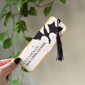 May include: A white bookmark with a black silhouette of a dragon and gold stars. The bookmark has a black tassel and the text "Save a dragon. Ride a wing leader."