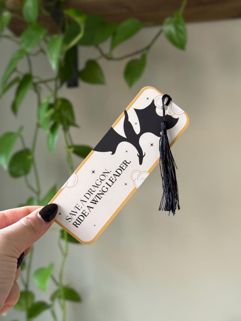 May include: A white bookmark with a black silhouette of a dragon and gold stars. The bookmark has a black tassel and the text "Save a dragon. Ride a wing leader."