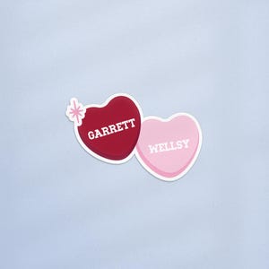 Könnte beinhalten: Zwei herzförmige Aufkleber. Einer ist tiefrot mit dem Namen "GARRETT" in Weiß. Der andere ist hellrosa mit "WELLSY" in Weiß. Ein kleines, weißes Sternmuster befindet sich auf dem roten Herzen. Die Aufkleber befinden sich auf hellblauem Hintergrund.