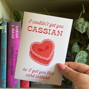May include: A white greeting card with red text and a heart-shaped cake illustration. The text reads "I couldn't get you Cassian, so I got you this card instead." The card is held in front of a bookshelf with books.