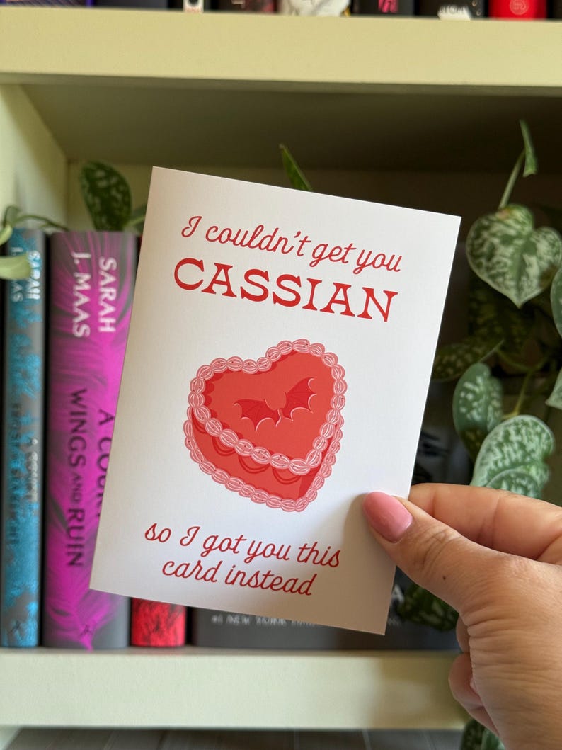 May include: A white greeting card with red text and a heart-shaped cake illustration. The text reads "I couldn't get you Cassian, so I got you this card instead." The card is held in front of a bookshelf with books.