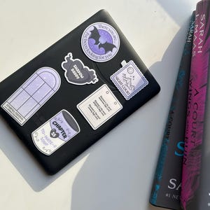 May include: A black e-reader with several stickers on it. The stickers are purple and white and feature a bat, a window, a soup can, a heart, and a stamp. The e-reader is sitting on a white surface.