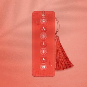 May include: Red bookmark with a tassel, featuring the name "CASSIAN" spelled out in white letters on red hearts. The bookmark has a white star design on either side of the name.