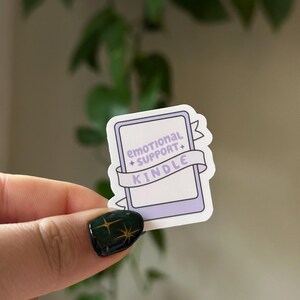 May include: A purple and white sticker with a Kindle e-reader design. The sticker has a banner that reads "emotional support + kindle".