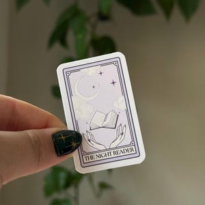 May include: A sticker with a purple background and white border. The sticker features a crescent moon, stars, and a book floating in the air. Two hands are reaching up to the book. The text "THE NIGHT READER" is at the bottom of the sticker.