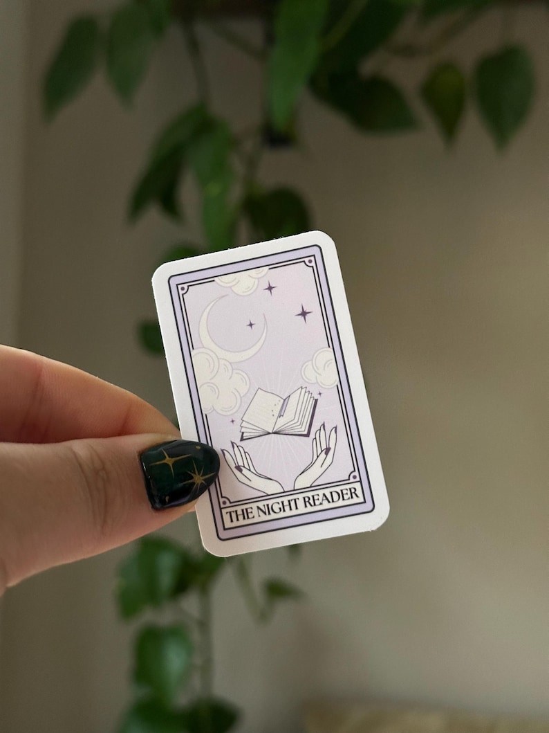 May include: A sticker with a purple background and white border. The sticker features a crescent moon, stars, and a book floating in the air. Two hands are reaching up to the book. The text "THE NIGHT READER" is at the bottom of the sticker.