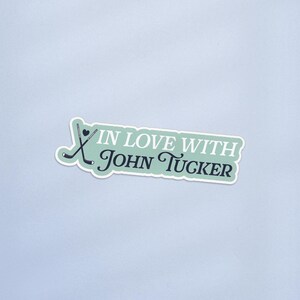 May include: A sticker with a light green background and white border. The sticker features two crossed hockey sticks and the text "IN LOVE WITH John Tucker" in a mix of fonts and colors.