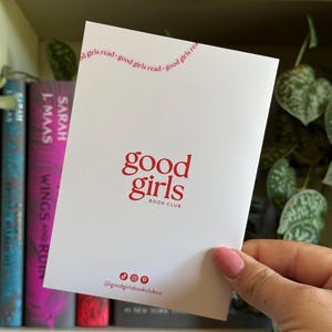 May include: A white card with the words "good girls" in red, with "BOOK CLUB" below. The top of the card reads "good girls read" in a curved line. The card is held in front of a bookshelf with books.