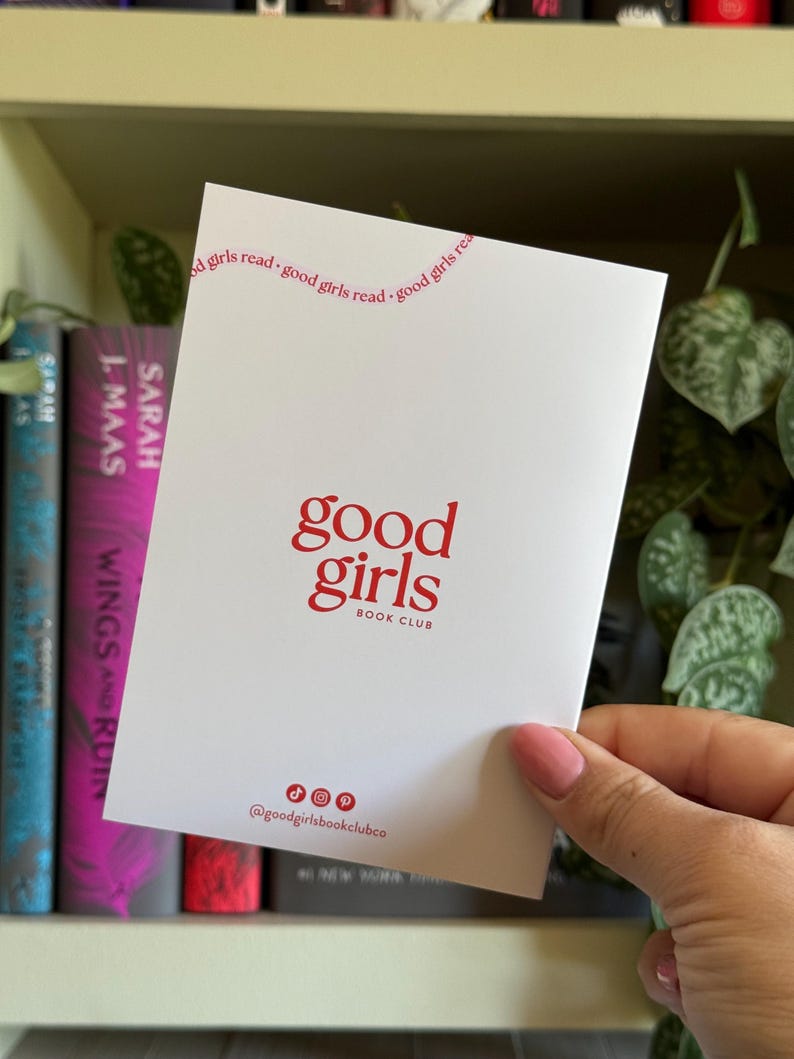 May include: A white card with the words "good girls" in red, with "BOOK CLUB" below. The top of the card reads "good girls read" in a curved line. The card is held in front of a bookshelf with books.