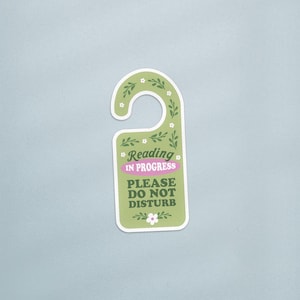 May include: A green door hanger with the text "Reading in progress PLEASE DO NOT DISTURB". The hanger has a floral design with white flowers and green leaves. The text "Reading in progress" is in a pink box.