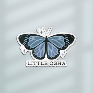 K&ouml;nnte beinhalten: Ein Aufkleber mit einem Schmetterlingsdesign mit schwarzen und blauen Fl&uuml;geln und einem wei&szlig;en Rand. Der Aufkleber hat den Text "LITTLE OSHA" in schwarzer Schrift. Dekorative Sternformen umgeben den Schmetterling.