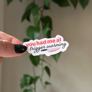 Może przedstawiać: Biała naklejka z tekstem "You had me at trigger warning" w czerwonych i czarnych literach. Obok tekstu narysowany jest czerwony n&oacute;ż z czarną rękojeścią.