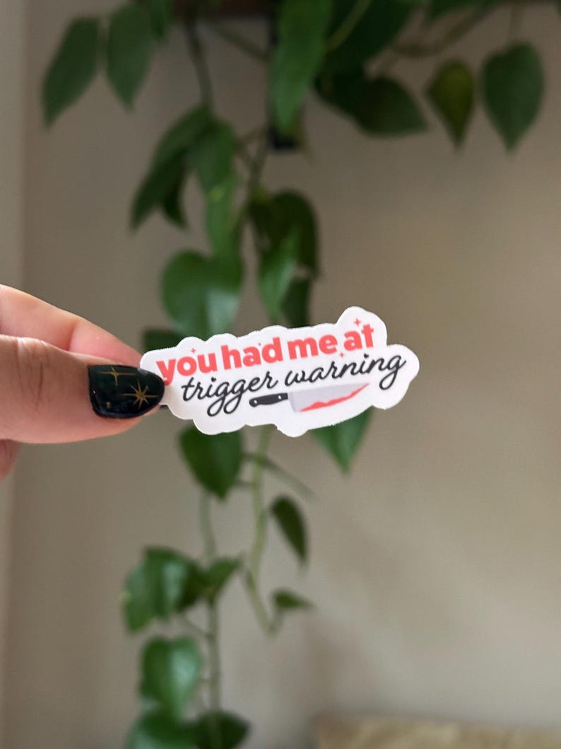 Może przedstawiać: Biała naklejka z tekstem "You had me at trigger warning" w czerwonych i czarnych literach. Obok tekstu narysowany jest czerwony n&oacute;ż z czarną rękojeścią.