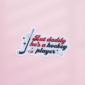 May include: A sticker with the text "But daddy he's a hockey player" in red and blue, with a hockey stick, puck, and hearts. The sticker has a light blue background and is on a pink background.