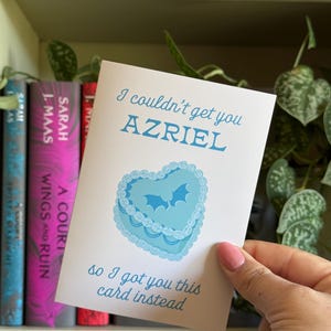 May include: A greeting card with the text "I couldn't get you AZRIEL, so I got you this card instead." The card features a blue heart-shaped cake design with bat silhouettes. Bookshelves are in the background.
