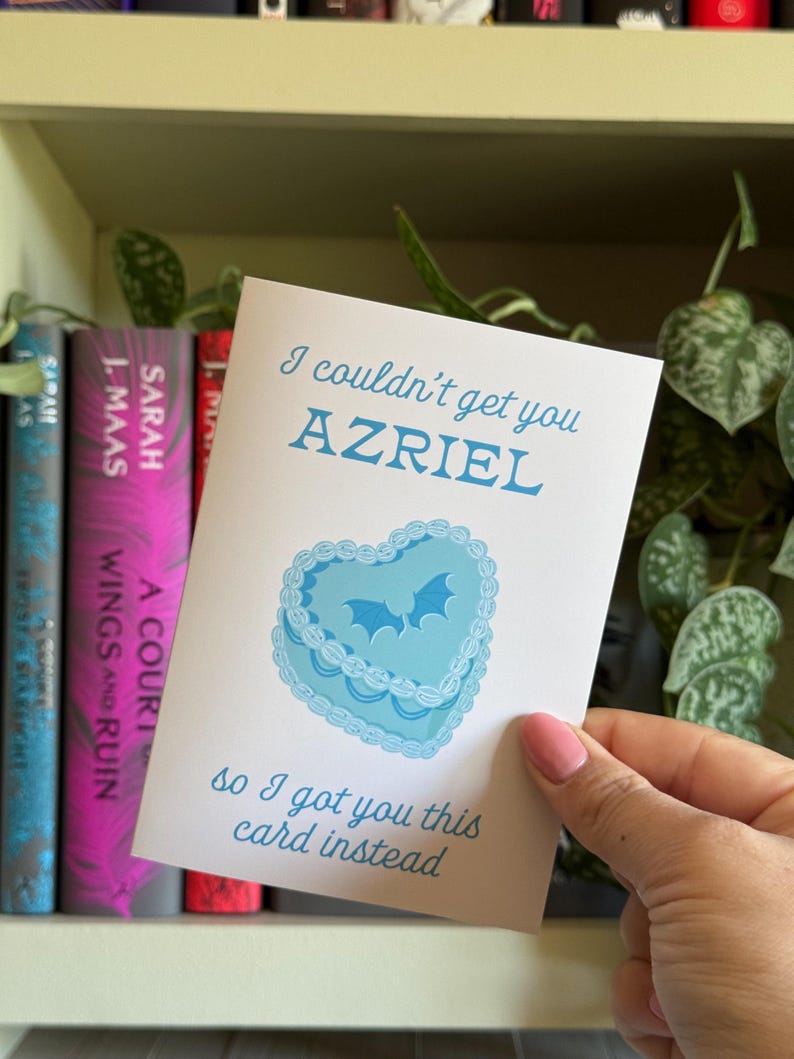 May include: A greeting card with the text "I couldn't get you AZRIEL, so I got you this card instead." The card features a blue heart-shaped cake design with bat silhouettes. Bookshelves are in the background.