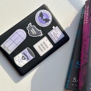 Op de afbeelding: Een zwarte e-reader met verschillende stickers erop. De stickers zijn paars en wit en bevatten een vleermuis, een raam, een blik soep, een hart en een postzegel. De e-reader ligt op een wit oppervlak.