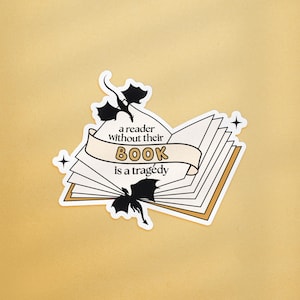Op de afbeelding: Een sticker met een open boek met de tekst "a reader without their BOOK is a tragedy". Twee zwarte draken silhouetten en sterren zijn ook opgenomen in het ontwerp. De sticker heeft een witte achtergrond met een gouden rand.