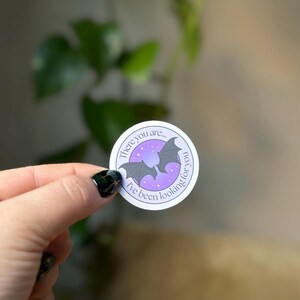 May include: Round sticker with a purple and white design featuring two gray bats and the text "There you are... I've been looking for you." The background is a gradient of purple and white.