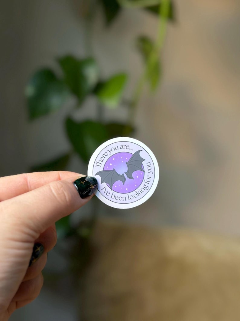 May include: Round sticker with a purple and white design featuring two gray bats and the text "There you are... I've been looking for you." The background is a gradient of purple and white.