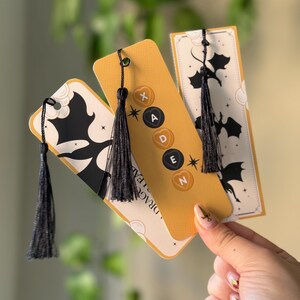 May include: Three yellow and white bookmark designs with black dragon silhouettes and black tassels. The bookmarks feature the text "Dragon Slayer" and black buttons with the letters "X", "A", "D", "E", and "N".