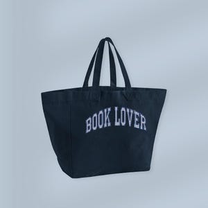 Peut inclure: Un sac fourre-tout bleu marine avec le texte "BOOK LOVER" en lettres blanches. Le sac a deux poignées et est fait d'un tissu robuste. Le texte a un motif à carreaux.
