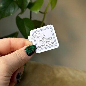 Op de afbeelding: Een witte sticker met een paarse rand en een stempelontwerp. De sticker toont een bergketen met een halve maan en sterren. De tekst "NIGHT COURT" is in zwart onder de bergen gedrukt.