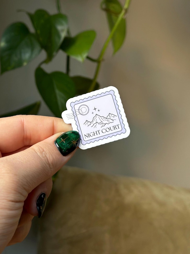 Op de afbeelding: Een witte sticker met een paarse rand en een stempelontwerp. De sticker toont een bergketen met een halve maan en sterren. De tekst "NIGHT COURT" is in zwart onder de bergen gedrukt.