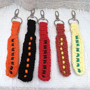 May include: Five crocheted keychains with beaded accents. The keychains are in various colors, including orange, black, red, brown, and yellow. Each keychain has a silver key ring.