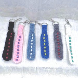 May include: A collection of six handmade crochet keychains in various colors. Each keychain features a bottle-shaped design with decorative beads. Colors include black, pink, blue, gray, and white. The keychains are attached to silver-tone metal clasps.