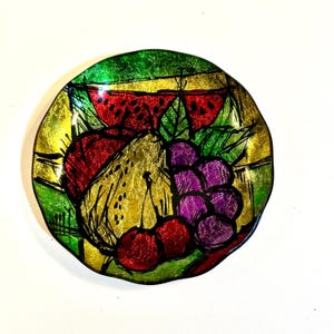 May include: A round, decorative glass plate with a fruit design. The plate features a watermelon, grapes, cherries, and a pear. The colors include red, green, yellow, and purple. The plate has a gold foil background and black outlines.