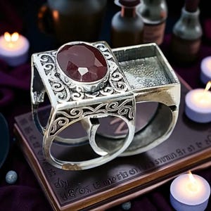May include: A silver ring with an ornate design and a large, oval, red gemstone. The ring has a hinged compartment. The ring is sitting on a book with text and surrounded by candles.