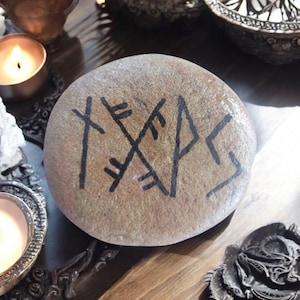 May include: A round, tan stone with black painted runes. The stone is smooth and appears to be natural. The runes are a stylized, geometric design. The stone is likely used for divination or spiritual practices.