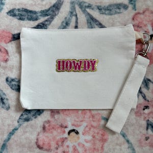 Puede incluir: Una bolsa de lona blanca con una correa para la muñeca. La bolsa presenta la palabra "HOWDY" bordada en rosa con ribete dorado. El fondo es un estampado floral en rosa, azul y blanco.
