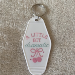 May include: White motel key chain with the text "A little bit dramatic" in pink and green lettering. The keychain features a pink bow and two pink cherries.