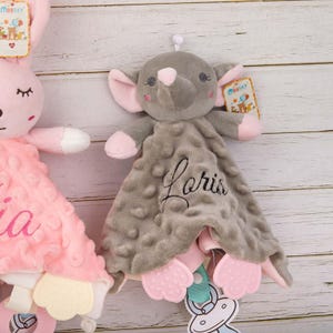 May include: A gray elephant-shaped baby security blanket with pink accents on the ears and feet. The blanket has a textured surface and the name "Paris" embroidered in cursive. A tag with the brand name "BBsky" is attached.