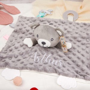 Peut inclure: Une couverture de sécurité grise pour bébé avec une tête et des bras d'ours en peluche. La couverture a une surface texturée et pointillée et le nom "Arthur" brodé en blanc. Comprend une sucette et un anneau de dentition.