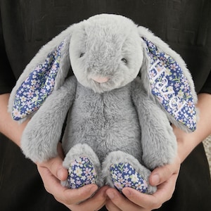 Puede incluir: Un peluche gris con forma de conejo, con orejas largas y una nariz rosa. El interior de las orejas y las plantas de los pies tienen un estampado floral azul. El conejo es suave y tierno, ideal para una colección de juguetes infantiles.