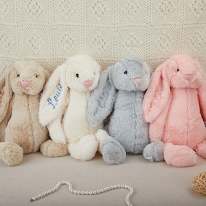 May include: Four soft toy bunny rabbits in beige, white, grey, and pink. Each has long floppy ears, a pink nose, and embroidered features. One bunny has the name "Louis" embroidered on its ear. The bunnies are designed to be cuddly.