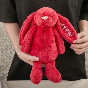 May include: A plush, red bunny rabbit with long floppy ears. The bunny has a pink nose and the name "Sophie" embroidered in white on one ear. The soft toy is held in front of a person wearing a black shirt.