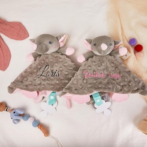 May include: Two gray elephant security blankets with pink accents and personalized names embroidered on the blanket. The blankets have a textured surface and are paired with a blue and white pacifier clip and a wooden teether.