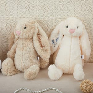 May include: Two plush bunny toys with long ears. The bunny on the left is beige with the name "Jonathan" embroidered on its ear. The bunny on the right is white with the name "Lou" embroidered on its ear.